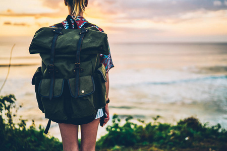年轻的女性旅行者穿着休闲服装，在日落时站在海岸线上欣赏无尽的海洋景观。带有背包的时髦女孩在户外放松的形象被剪掉了