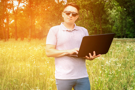 休闲年轻人戴着太阳镜，手提电脑站在户外草地上，输入自由和幸福的概念。 年轻人在阳光明媚的日子里用笔记本电脑在大自然中放松。 穿灰