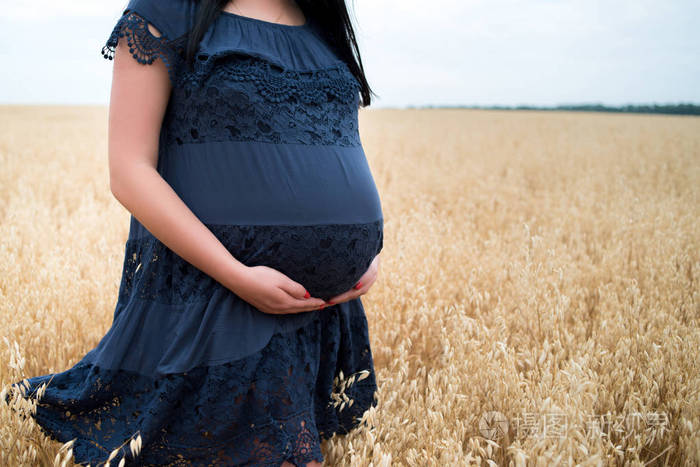 年轻漂亮的9个月孕妇穿着蓝色连衣裙,抱着肚子,在夏日户外的燕麦田里