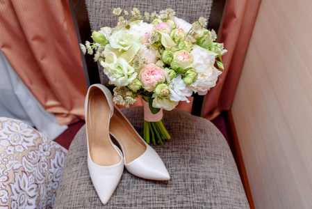 在室内的椅子上合上漂亮的粉红色和白色玫瑰和新娘鞋的婚礼花束。 婚礼的早晨准备。 结婚新娘配饰。 新娘花束和鞋子