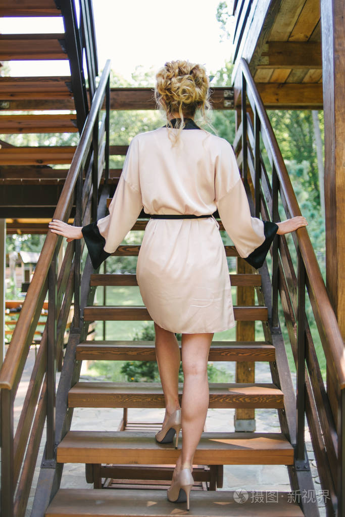 年轻女子穿着丝绸长袍穿着带有卷曲发型的高跟鞋走在户外的木制楼梯上