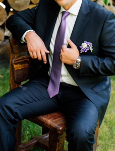 时髦的新郎穿着黑色西装领带和户外的布托尼埃。 新郎坐在椅子上，拿着夹克的领子