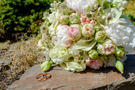 两个金色的结婚戒指在石头上，美丽的婚礼花束白色和粉红色的牡丹和玫瑰户外选择性的集中