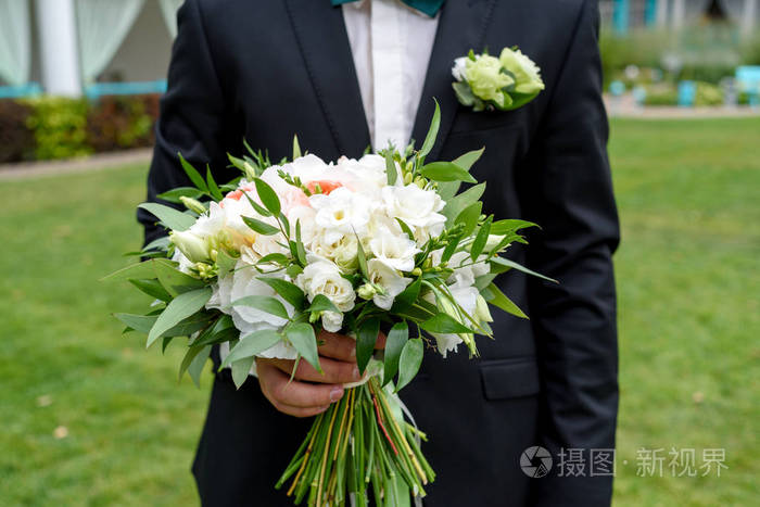 新娘婚礼的花束是白色的花和绿色的植物，新郎手里拿着丝带。 新郎在户外等新娘。 年轻英俊的新郎，穿着黑色西装，站着，拿着结婚花束