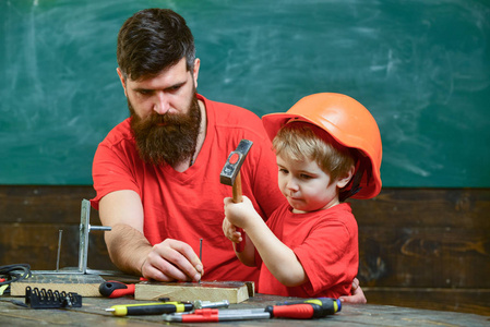 男孩, 孩子忙着保护头盔学习用锤子和爸爸在一起。小助手的概念。父亲用胡子教小儿子使用工具, 锤击, 黑板在背景上