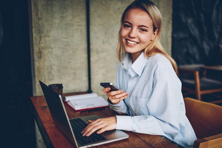 成功的女性自由职业者在使用4G互联网在笔记本电脑设备工作时微笑的肖像。积极的博主在更新上网本软件的同时，在智能手机上发送短信