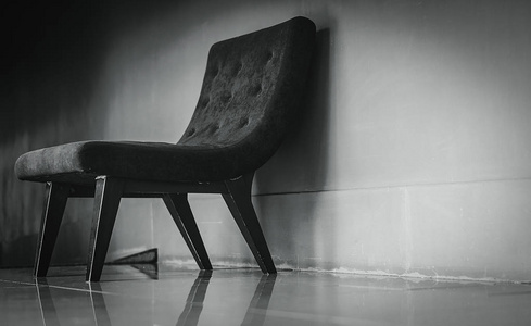 黑色经典椅子，独特的设计在空休息室附近的混凝土墙上，黑暗和戏剧性的背景。 沮丧悲伤和孤独的概念。 客厅里有一张空扶手椅。 室内