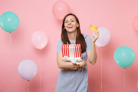 快乐的年轻女子的肖像，穿着蓝色连衣裙，拿着信用卡和红色盒子，礼物呈现在彩色气球的粉彩背景上。 例如生日聚会真诚的情感