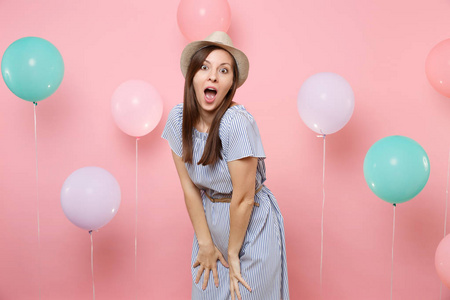 震惊的肖像，惊讶的年轻女子，戴着稻草夏帽和蓝色连衣裙，站在彩色气球的粉红色背景上。 生日聚会真诚的情感观念