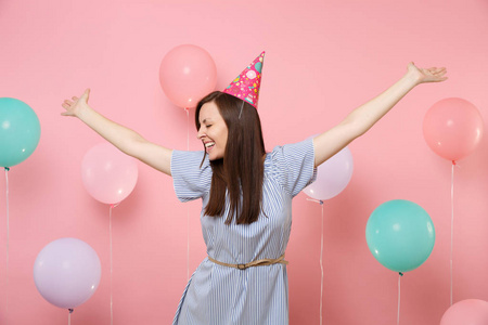 笑的年轻女人的肖像，闭着眼睛戴着生日帽和蓝色连衣裙，双手在粉红色的背景上展开，五颜六色的空气包。 生日聚会真诚的情感观念