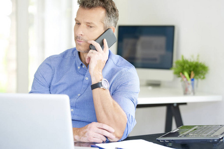 中年商人的肖像，穿着衬衫，坐在办公室工作。 专业人士使用他的手机，在笔记本电脑上工作时与某人交谈。 多任务处理。