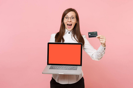 年轻震惊的商界女性戴着眼镜，手持信用卡笔记本电脑，电脑上有空白的空屏幕，孤立在粉红色的背景上。 女老板。 成就事业财富。 广告复