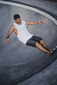 一个穿着白色T恤和帽子的时髦年轻人在游泳池里溜冰的溜冰场上玩滑板。