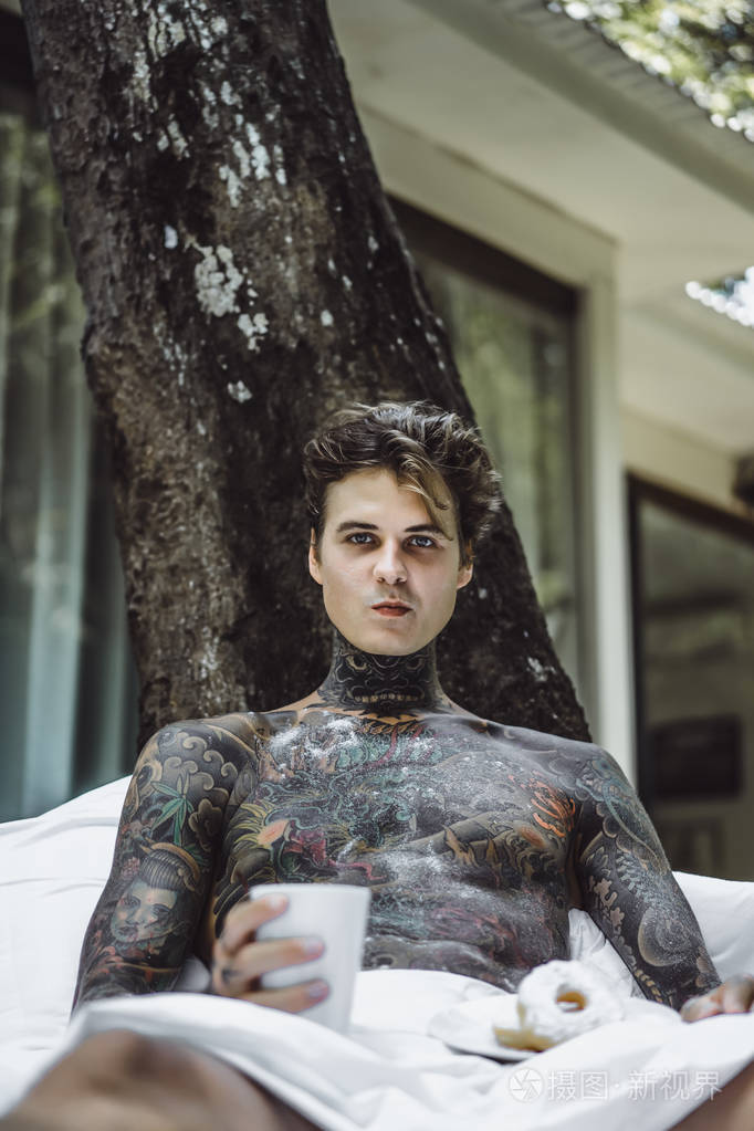 年轻英俊的纹身男在户外露天的床上吃早餐