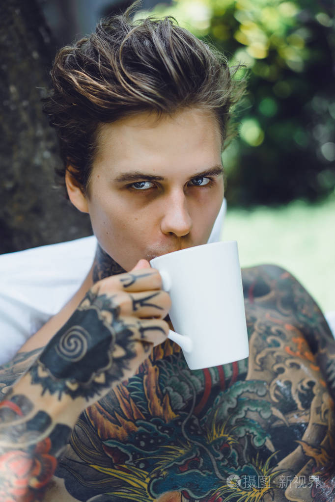 年轻英俊的纹身男在户外露天的床上吃早餐