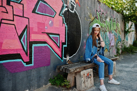 时髦的休闲时髦女孩戴着帽子和牛仔裤，戴着手机耳机听音乐，对着大的涂鸦墙和。