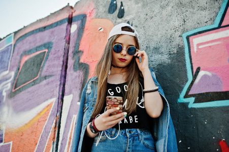 时尚的休闲时髦女孩戴着帽子太阳镜和牛仔裤，戴着手机耳机听音乐，对着大的涂鸦墙。