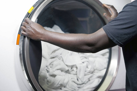 客栈里一名非洲男性洗衣工人的手从洗衣机里拿出一条干净的毛巾。 时尚酒店洗衣房的洗衣机。 在酒店洗衣服。