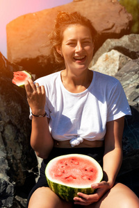 年轻漂亮的女人开心地笑着，笑着，两腿之间抱着一个大西瓜，吃着东西，在海边的一个夏天坐在石头上