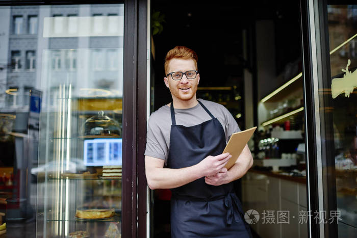 面包店老板的肖像他拿着菜单邀请去购物穿围裙的积极服务员微笑着看着