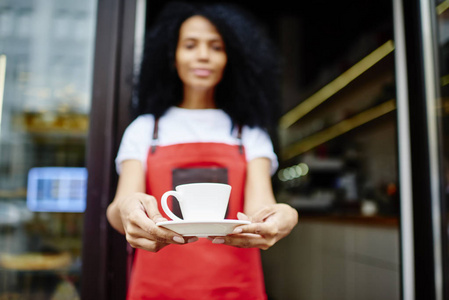 有选择地专注于非裔美国人女咖啡师手中的白杯香气咖啡饮料。背景模糊的非裔美国人女服务员穿着红色围裙提供自己的饮料