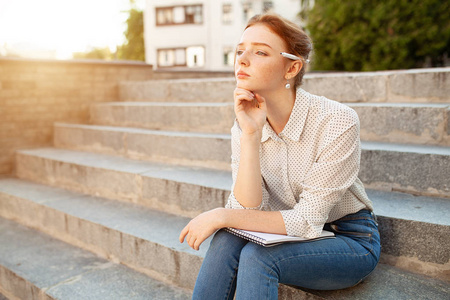 美丽的年轻的红发女孩与雀斑坐在楼梯上, 并在笔记本上做笔记。学生的肖像。远程教育。作业