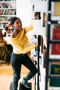 美丽的非裔美国嬉皮士学生在图书馆书架上挑选文学书籍时，微笑着看着相机的肖像。在公共书店图书馆工作的图书管理员