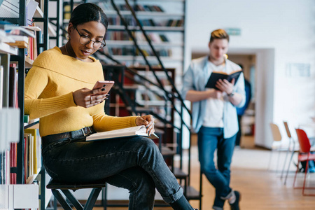 非裔美国人在现代智能手机网站上的阅读新闻，使用无线互联网连接，坐在图书馆里。深色皮肤的学生更新手机，而时髦的家伙在后台阅读书籍