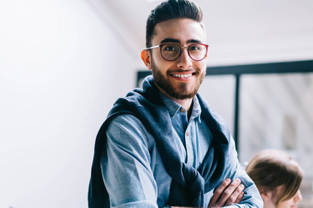 成功的年轻企业家戴着眼镜进行视力矫正的半长画像，微笑着面对镜头。穿着正式服装的积极商人和站在现代办公室的交叉手