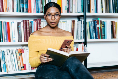 美丽的非裔美国年轻妇女的肖像，戴着时尚的眼镜，用于视力矫正，看着相机，同时更新手机上的应用程序，并坐在图书馆的书架旁，使用公共互