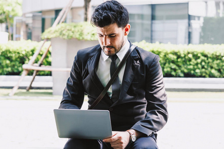 英俊的商人穿着黑色优雅的西装，坐在城市里用笔记本电脑工作。商业和创业理念概念