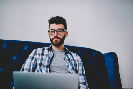 年轻的白种人时髦的戴着时髦眼镜的时髦帅哥坐在舒适的沙发上，拿着上网本，看着相机，假扮着笔记本电脑的假扮