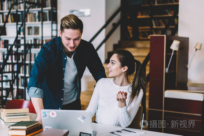 男女学生在合作做作业时面带微笑地用笔记本电脑交谈,积极的时髦女孩
