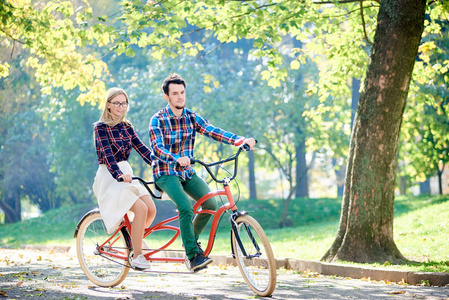 年轻活跃的微笑旅行者夫妇英俊的胡须男人和迷人的金发女人一起骑着双人自行车沿着小径，在明亮的阳光下，美丽的公园在高大的树木下。