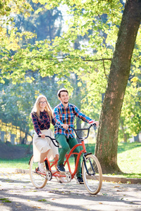 年轻的活跃的快乐的旅行者夫妇英俊的留着胡须的男人和迷人的金发女人，早上在高树下的秋天公园里，一起骑着双人自行车沿着小路。