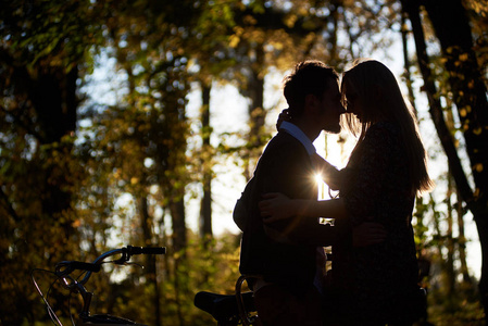 在黑暗的秋天公园或森林中，在模糊的金色背景下，由夕阳的轮廓照亮的浪漫夫妇年轻人和迷人的女孩在户外的串联自行车上亲密无间。