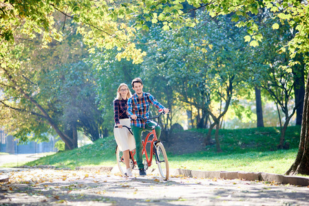年轻活跃的微笑旅行者夫妇英俊的胡须男人和迷人的金发女人一起骑着双人自行车沿着噼啪作响的小路在明亮的早晨阳光下美丽的公园树下。