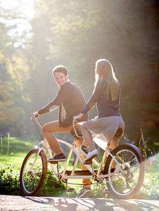 年轻的幸福夫妇留着胡须，微笑着的男人和金发的有吸引力的女人在夏日公园或森林里骑着双人自行车，在模糊的阳光明媚的绿色树木树叶背景下