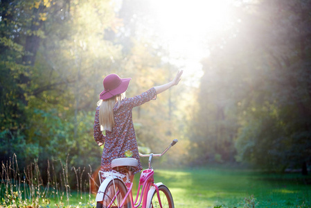 在铺好的夏日公园小巷里，骑着一辆豪华自行车，欣赏美丽的傍晚日落，她的后景是苗条的金发，时尚的长头发，漂亮的女孩，穿着短裙子，戴着