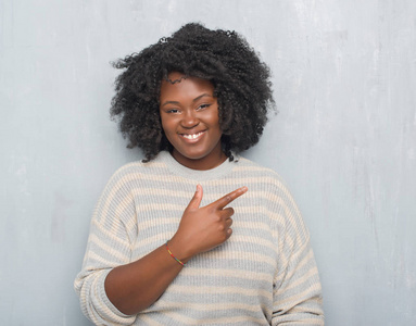 年轻的非裔美国人加码的女人，穿着灰色的灰色灰色墙壁，穿着一件毛衣，欢快地微笑着，用手和手指指向一边，脸上洋溢着快乐和自然的表情，