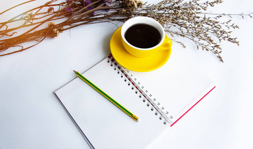 黄色的陶瓷咖啡杯和铅笔放在打开的书柜旁的干花白色桌子上