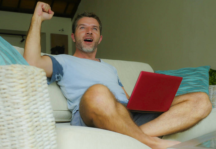 家庭生活方式室内照片年轻有魅力和快乐的男人坐在家里沙发上放松，使用互联网，在笔记本电脑上工作，表示胜利，作为胜利者，兴奋和快乐