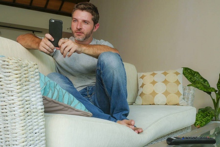 家庭生活方式室内肖像年轻英俊有吸引力的快乐男人使用互联网约会在线应用程序在手机上或网络坐在家里沙发沙发与智能手机轻松。