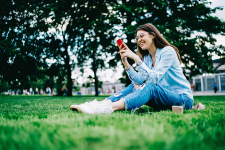 快乐的年轻女子戴着眼镜，笑着看着有趣的视频，在公园休闲时浏览着手机，微笑的时髦女孩坐在绿草上，拿着手机，在网上和朋友聊天