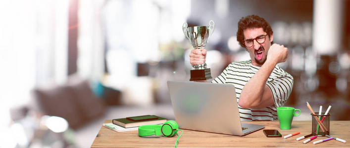 年轻的疯狂平面设计师在办公桌上拿着笔记本电脑和奖杯