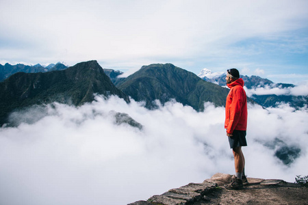 有经验的男性游客穿着活动服装，享受着平静和灵感，高山覆盖着白雾，站在山顶的边缘。年轻的旅行者欣赏着令人叹为观止的景色