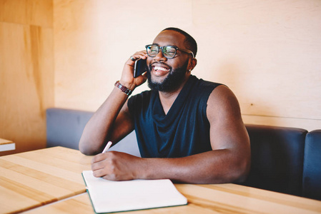 快乐的非裔美国人嬉皮士在与朋友的电话交谈中大笑，写着记事本，快乐的年轻，深色皮肤的男人，满足于得到便宜的电话费