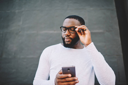 年轻的深色皮肤的嬉皮士戴着眼镜，通过智能手机阅读收入信息，站在户外，时髦的穿着非裔美国人博主用手机通过4g观看视频