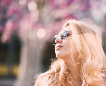 在阳光明媚的日子里，美丽的年轻金发女孩戴着太阳镜，嘴唇蓬松，的女性身材摆在街上，背景是粉红色的花