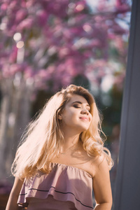 在阳光明媚的日子里，美丽的年轻金发女孩戴着太阳镜，嘴唇蓬松，的女性身材摆在街上，背景是粉红色的花